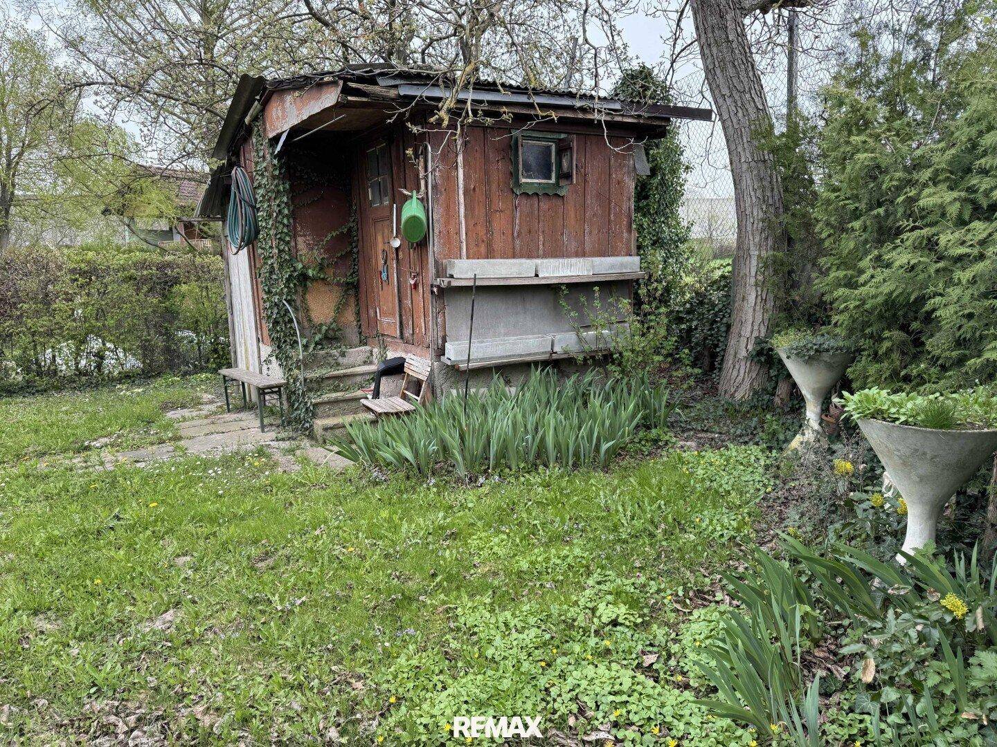 Rustikales Gartenhaus aus Holz mit kleiner Veranda, umgeben von Grünflächen und Pflanzen.
