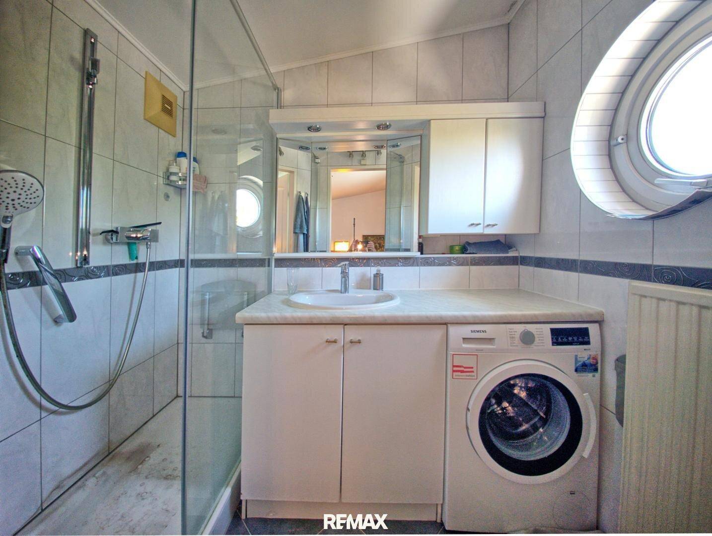 Helles Badezimmer mit Dusche, Waschbecken, Spiegelschrank und integrierter Waschmaschine.