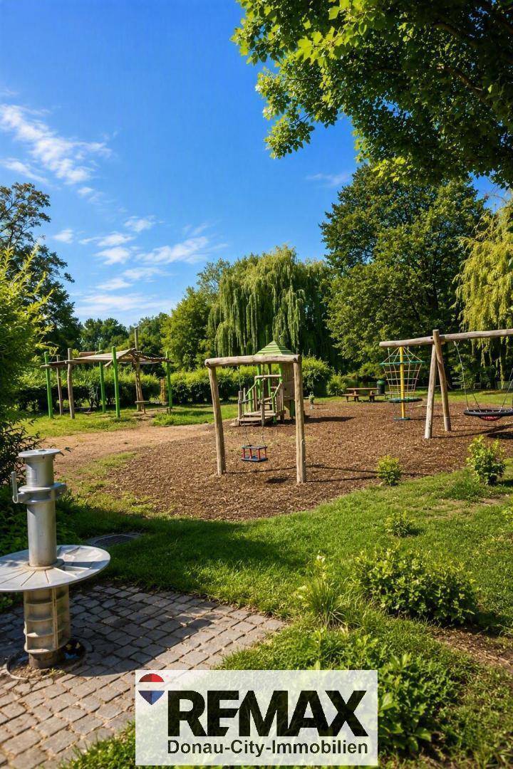 Großer Kinderspielplatz mit Schaukeln und Spielgeräten, umgeben von schattenspendenden Bäumen.