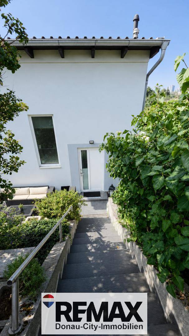 Moderner Hauseingang mit Treppe und Geländer, umgeben von grüner Bepflanzung.