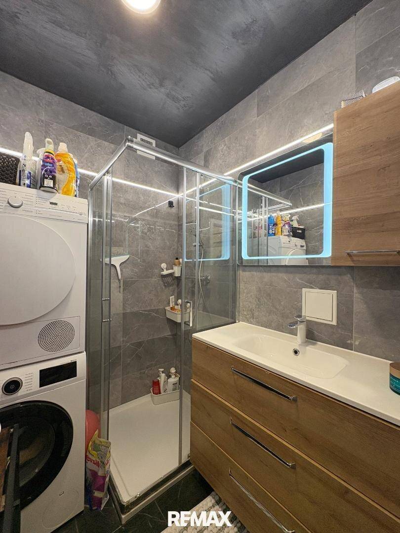Modernes Badezimmer mit grauen Fliesen, Duschkabine, Waschbecken und Waschmaschine.