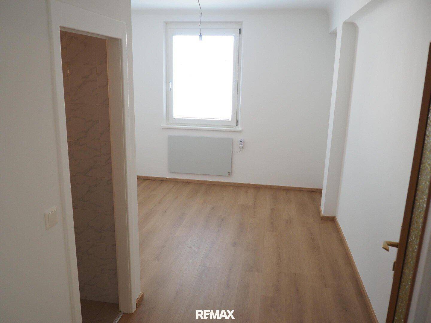 Helles Zimmer mit Laminatboden und Fenster, angrenzend an einen gefliesten Bereich.