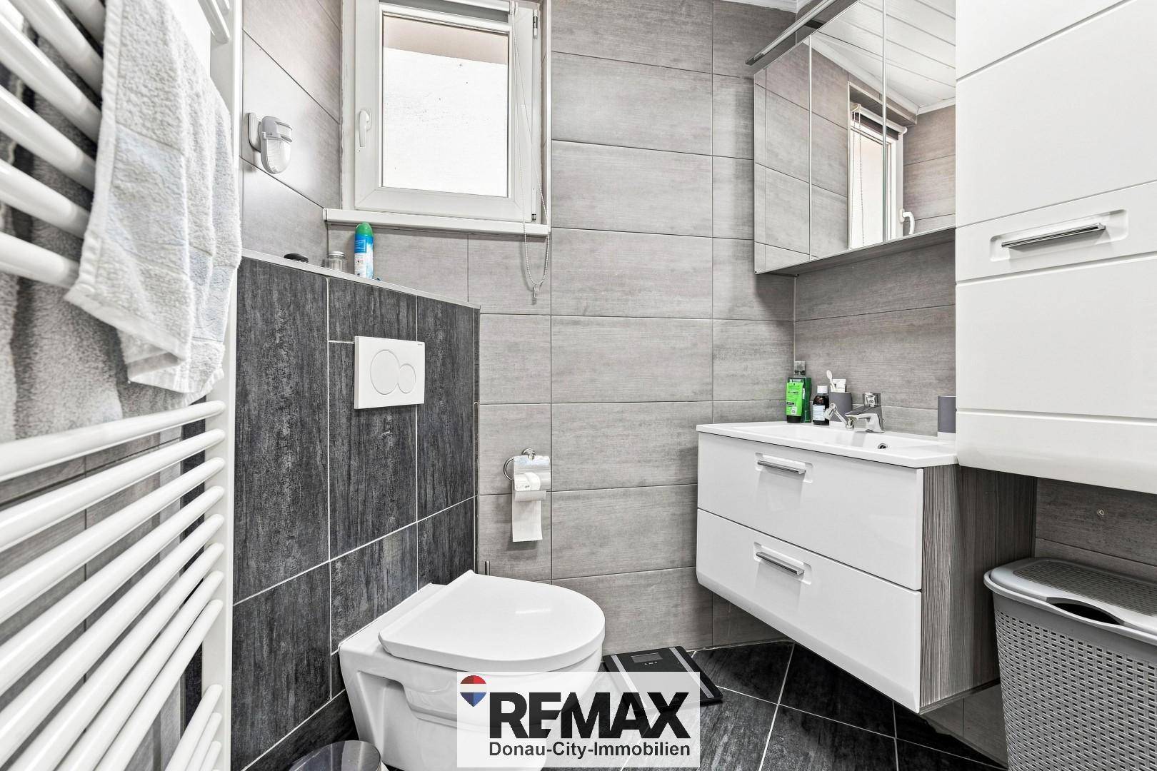 Modernes Badezimmer mit grauen Fliesen, wandhängendem WC und Waschtischunterschrank.