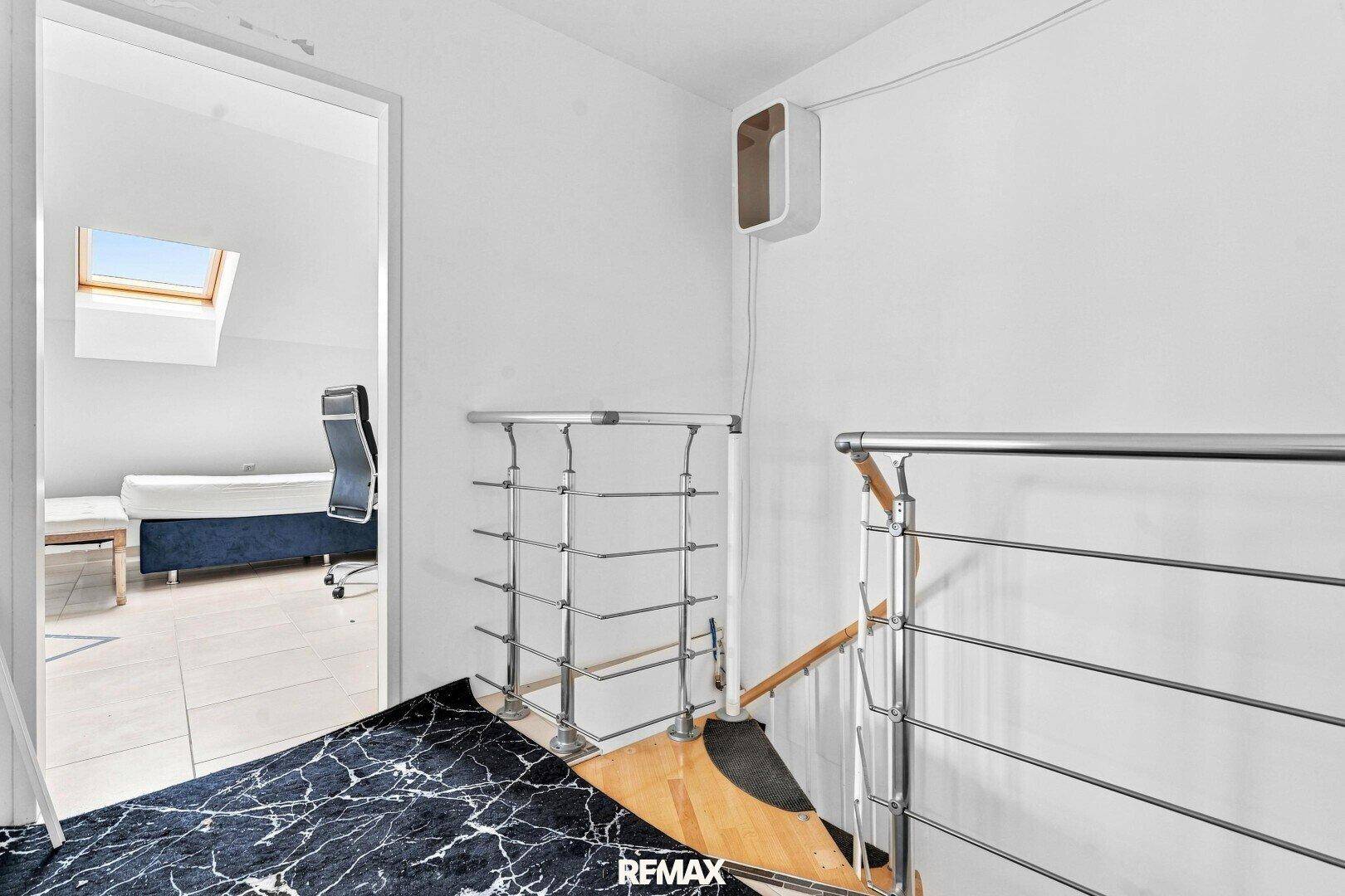 Treppenabsatz mit Metallgeländer, der zu einem hellen Dachzimmer mit Dachfenster führt.