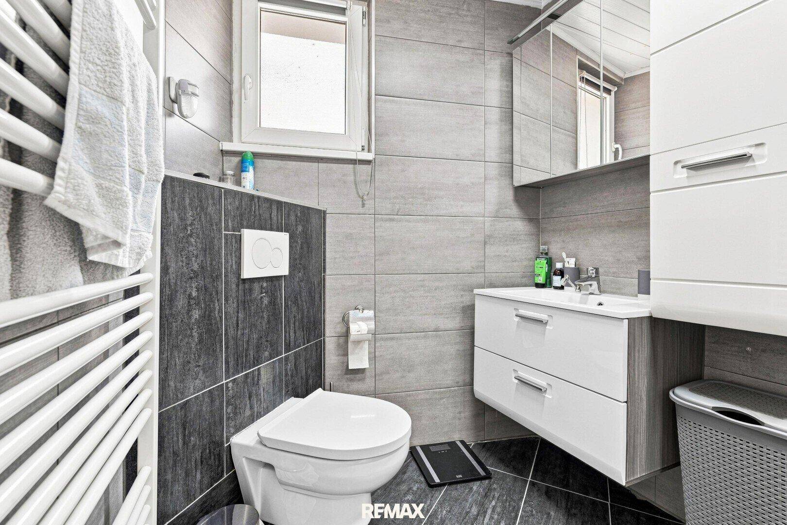 Modernes Badezimmer mit grauen Fliesen, wandhängendem WC, Waschbecken und beheiztem Handtuchhalter.