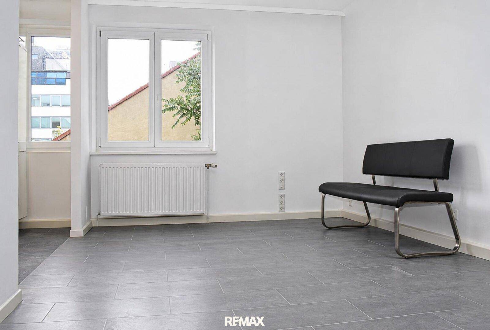 Heller, leerer Raum mit großem Fenster, Heizkörper und grauem Fliesenboden.