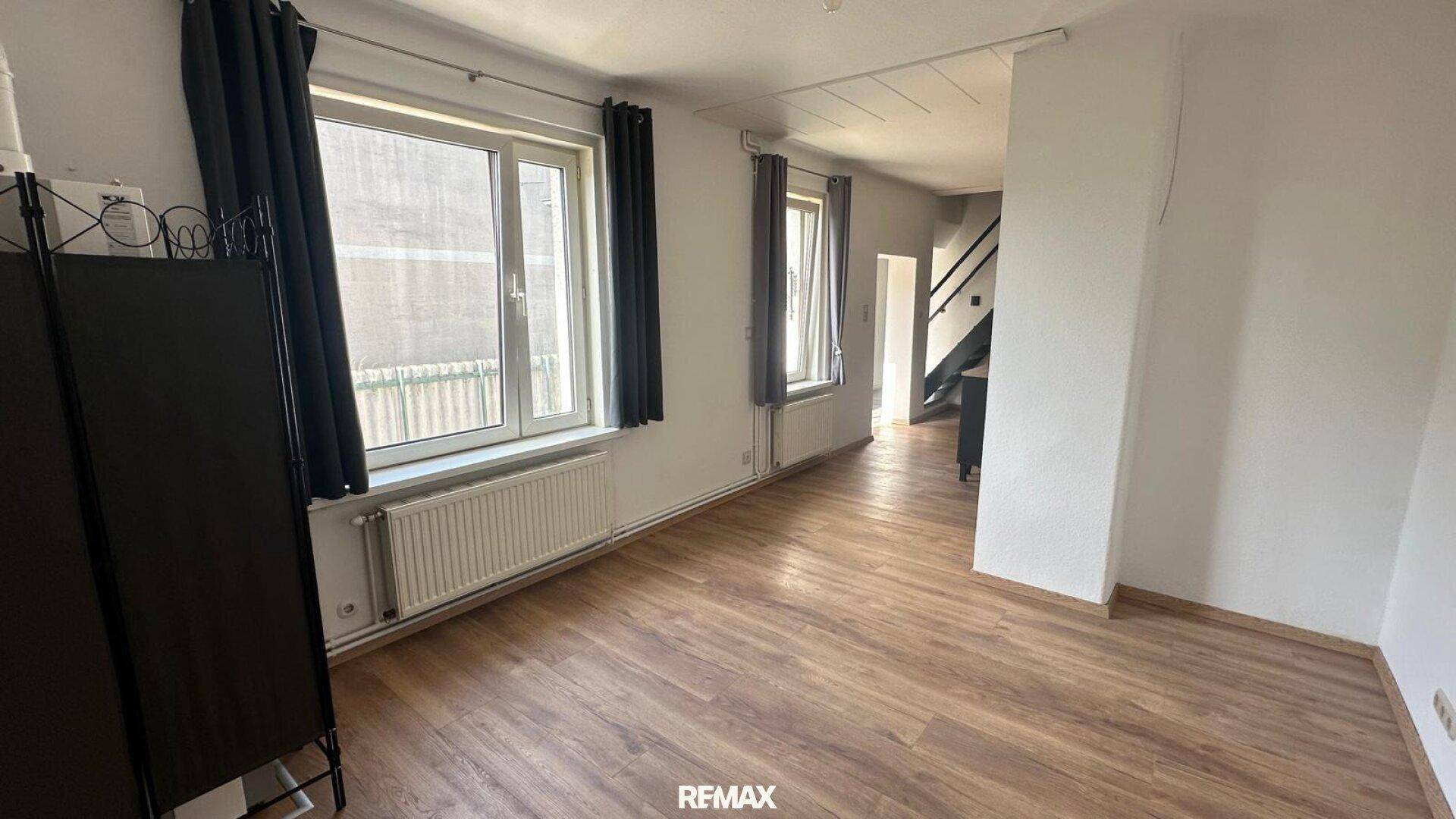 Geräumiges Wohnzimmer mit Laminatboden, großen Fenstern und Zugang zur Treppe.