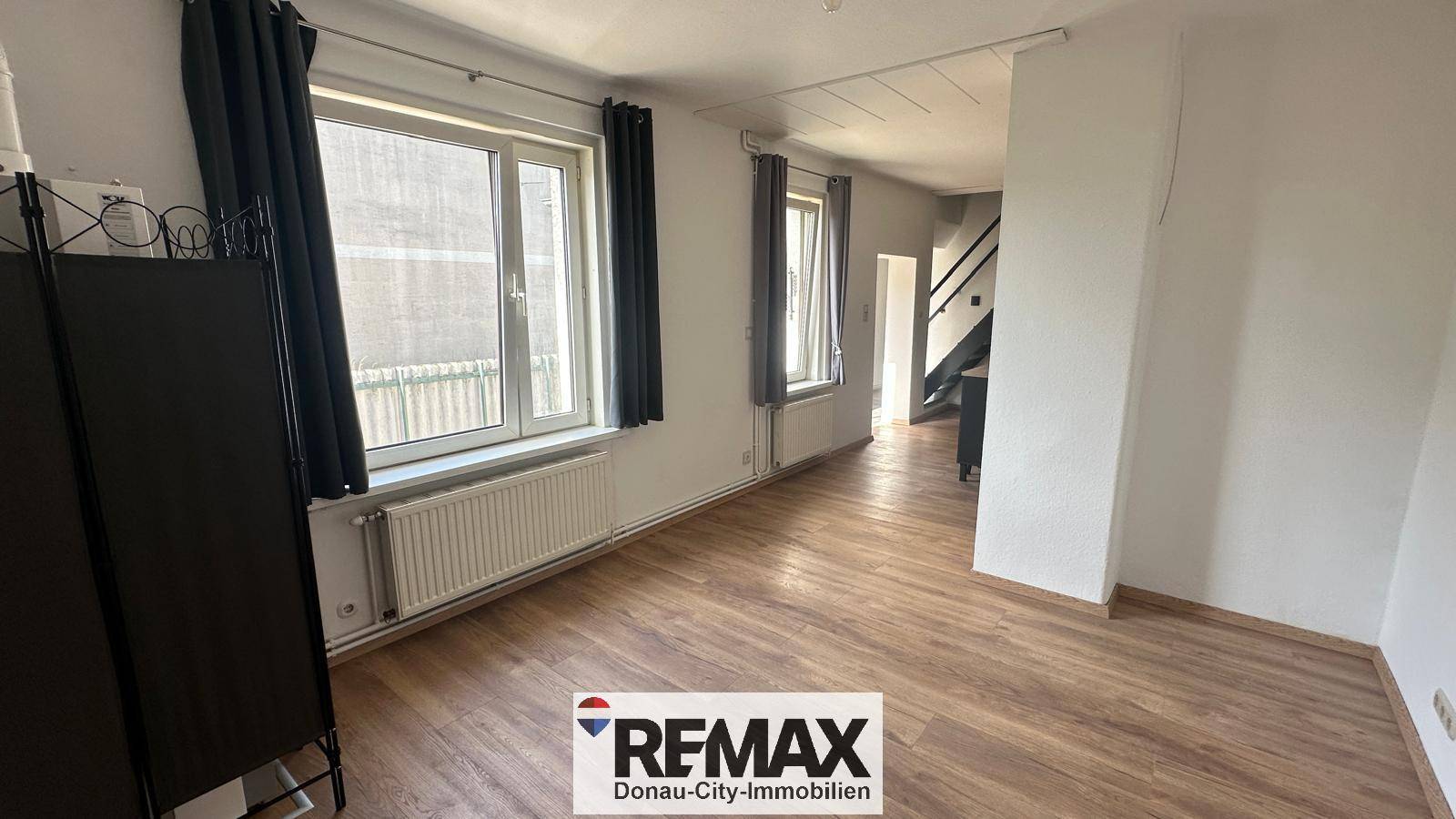 Helles Wohnzimmer mit Laminatboden, zwei Fenstern und dunklen Vorhängen.