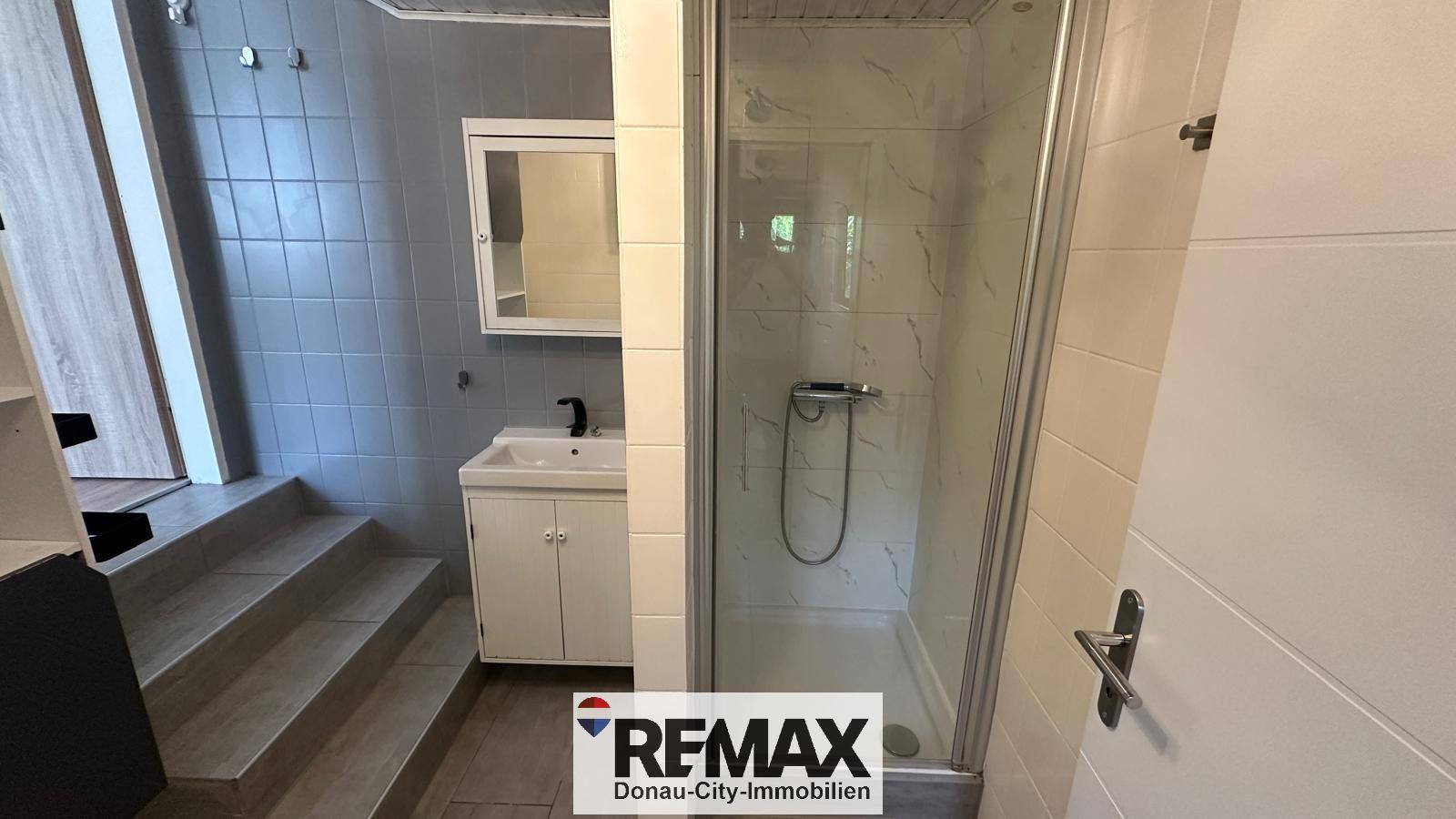 Badezimmer mit Duschkabine, Waschbecken, Spiegelschrank und grauen Fliesen.
