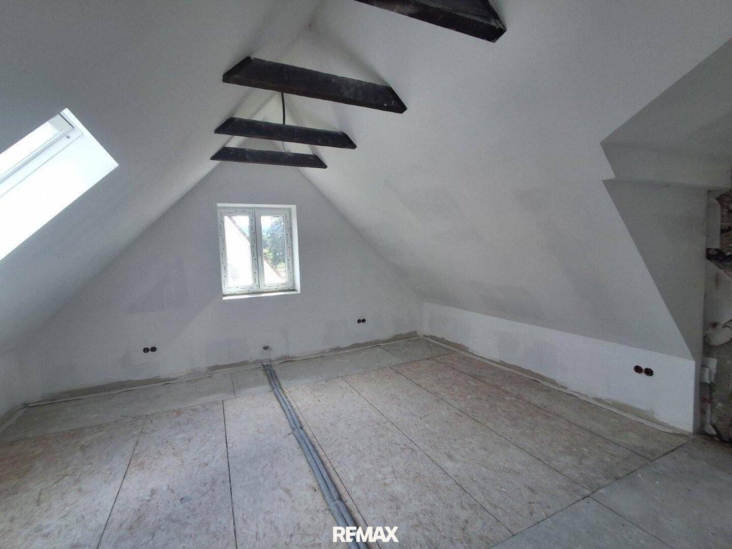 Dachgeschossraum im Ausbau mit schrägen Decken, sichtbaren Holzbalken und einem Dachfenster.