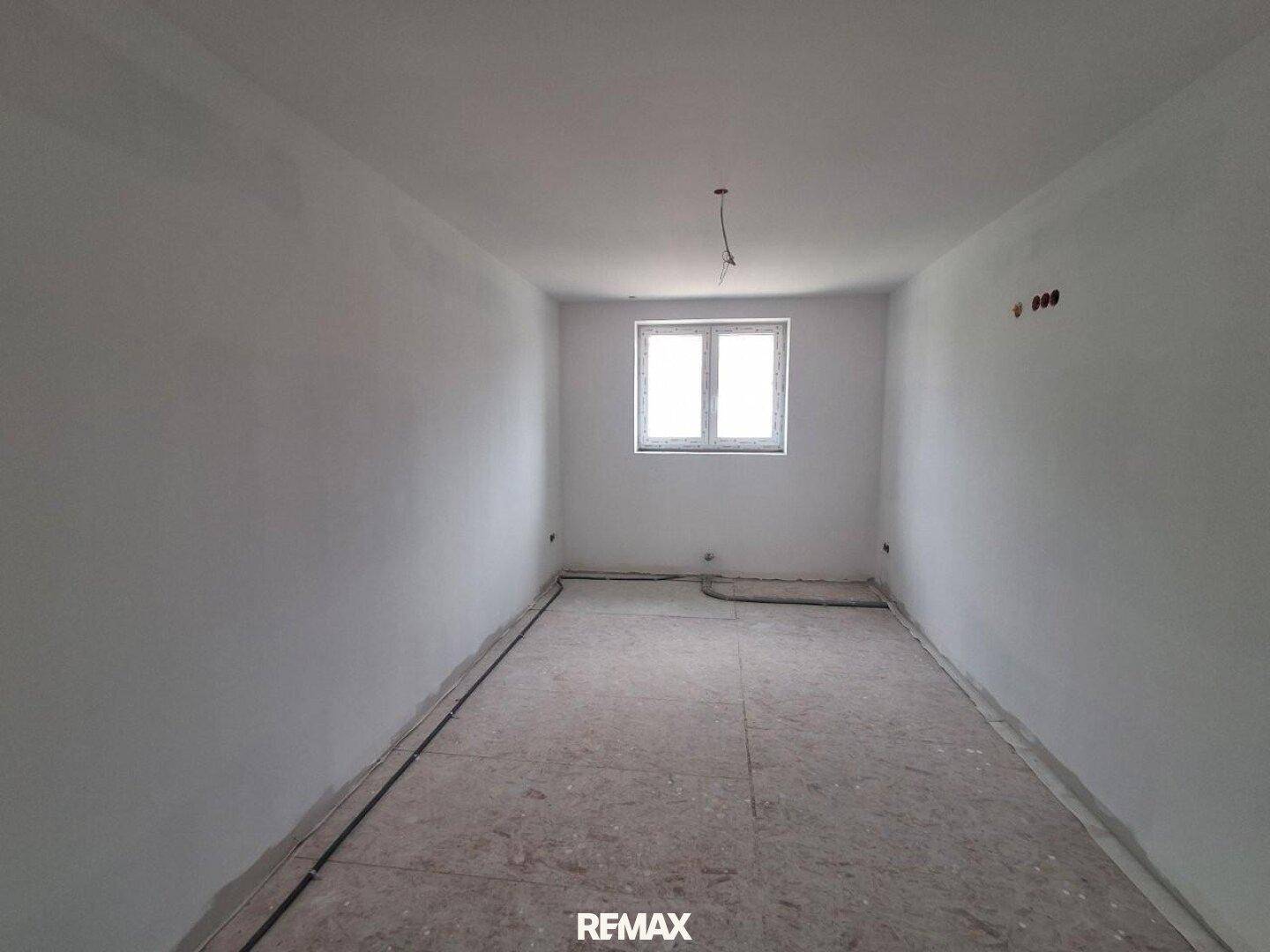 Leerer Raum im Rohbau mit weiß verputzten Wänden und einem Fenster, das Tageslicht spendet.
