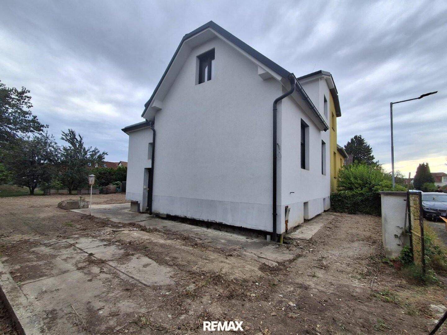 Seitenansicht des renovierten Hauses mit weißer Fassade und ungestaltetem Außenbereich.