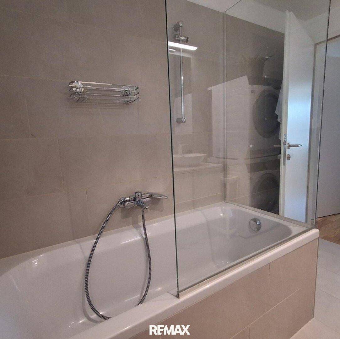 Badezimmer mit Badewanne und Duschfunktion, durch eine Glaswand abgetrennt, hell gefliest.