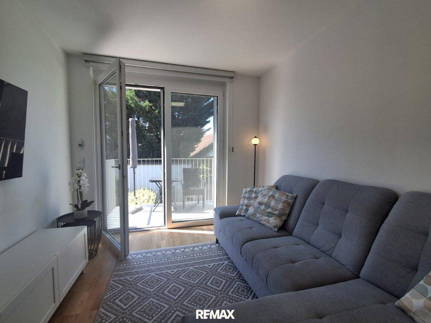 Gemütliches Wohnzimmer mit grauem Sofa, Teppich und Zugang zu einem sonnigen Balkon.