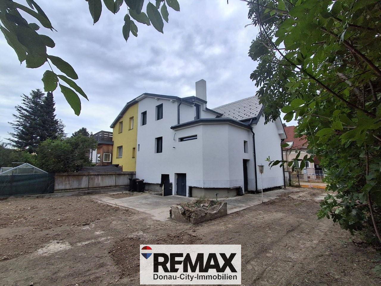 Rückansicht des Hauses mit weißer Fassade und unfertigem Gartenbereich, umgeben von Bäumen.