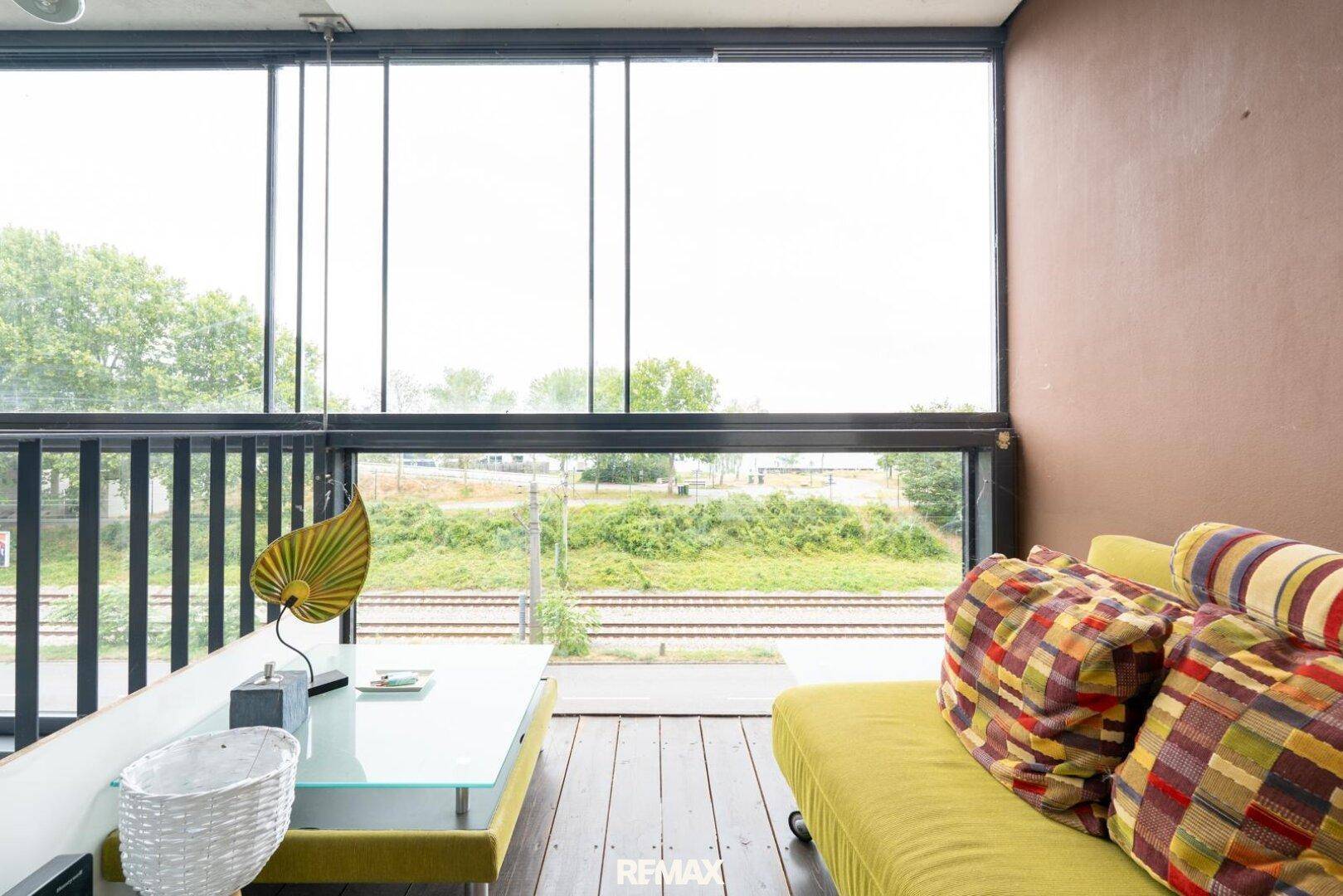 Heller Wohnbereich mit großen Fenstern und Blick auf Bahngleise und Grünflächen.