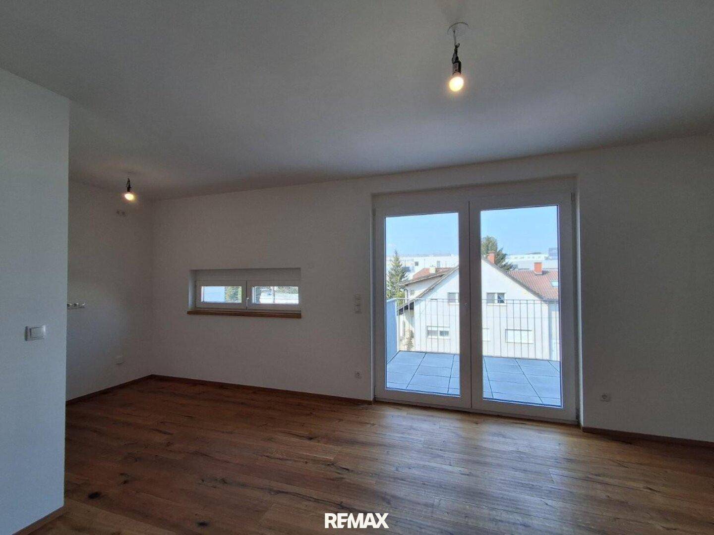 Heller, unmöblierter Raum mit Holzboden und großen Fenstern zum Außenbereich.
