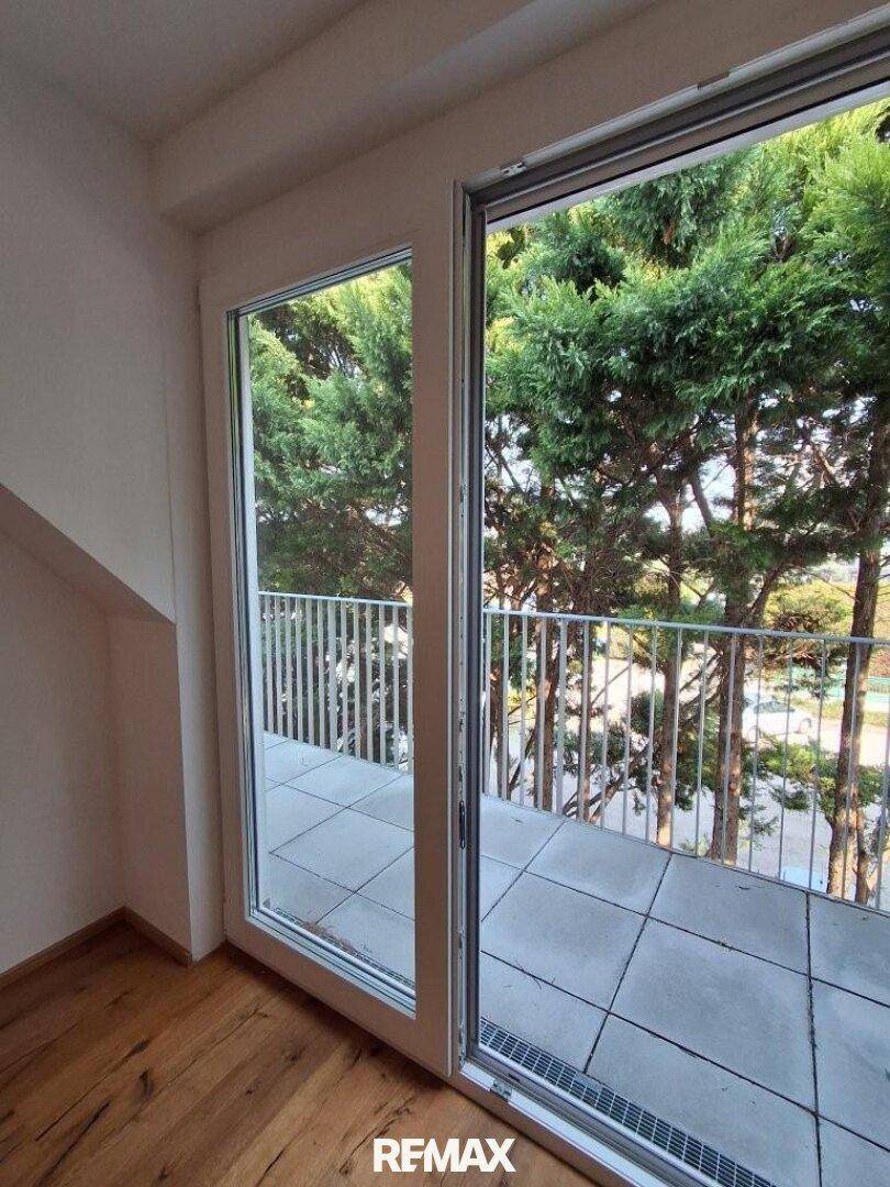 Heller Raum mit Holzboden und großem Fenster zum Balkon mit Grünblick.