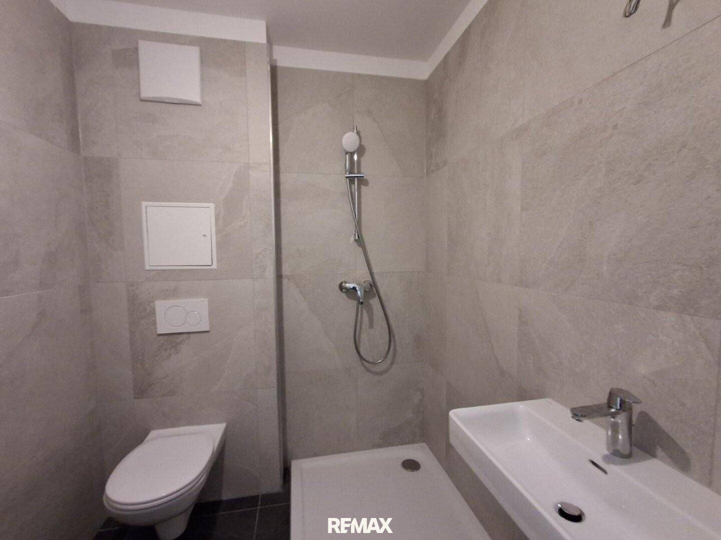 Modernes Badezimmer mit grauen Fliesen, Dusche und wandhängendem WC.