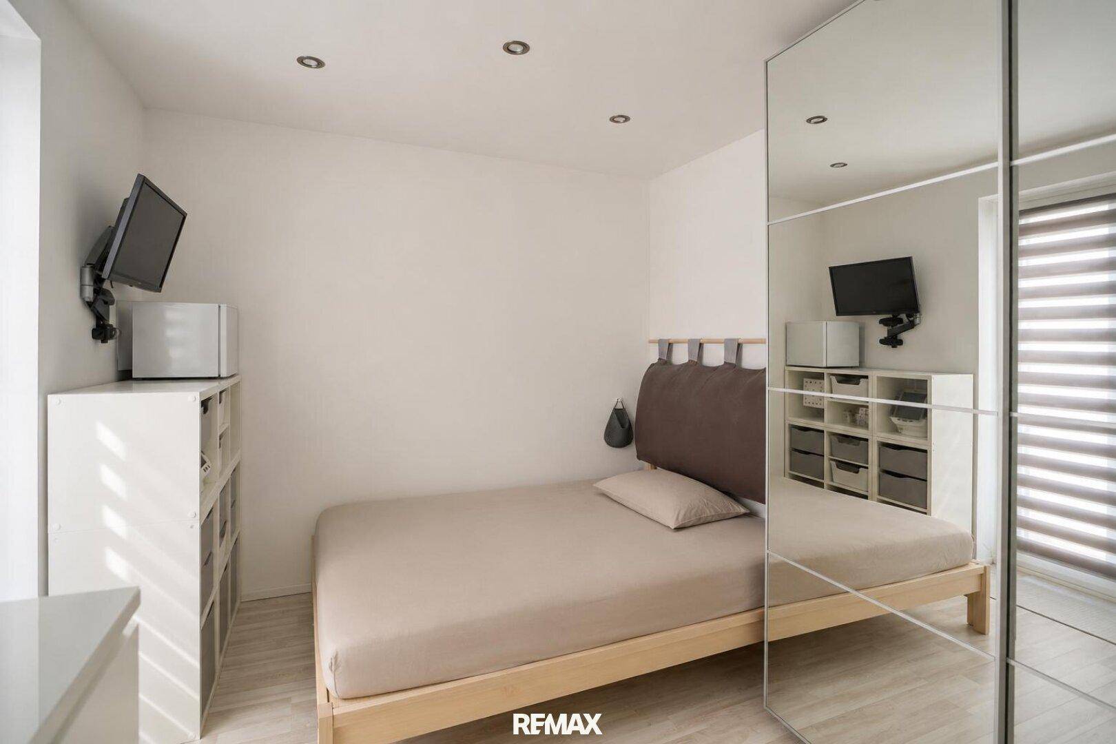 Helles Schlafzimmer mit Bett, Spiegelschrank und Fernseher an der Wand.