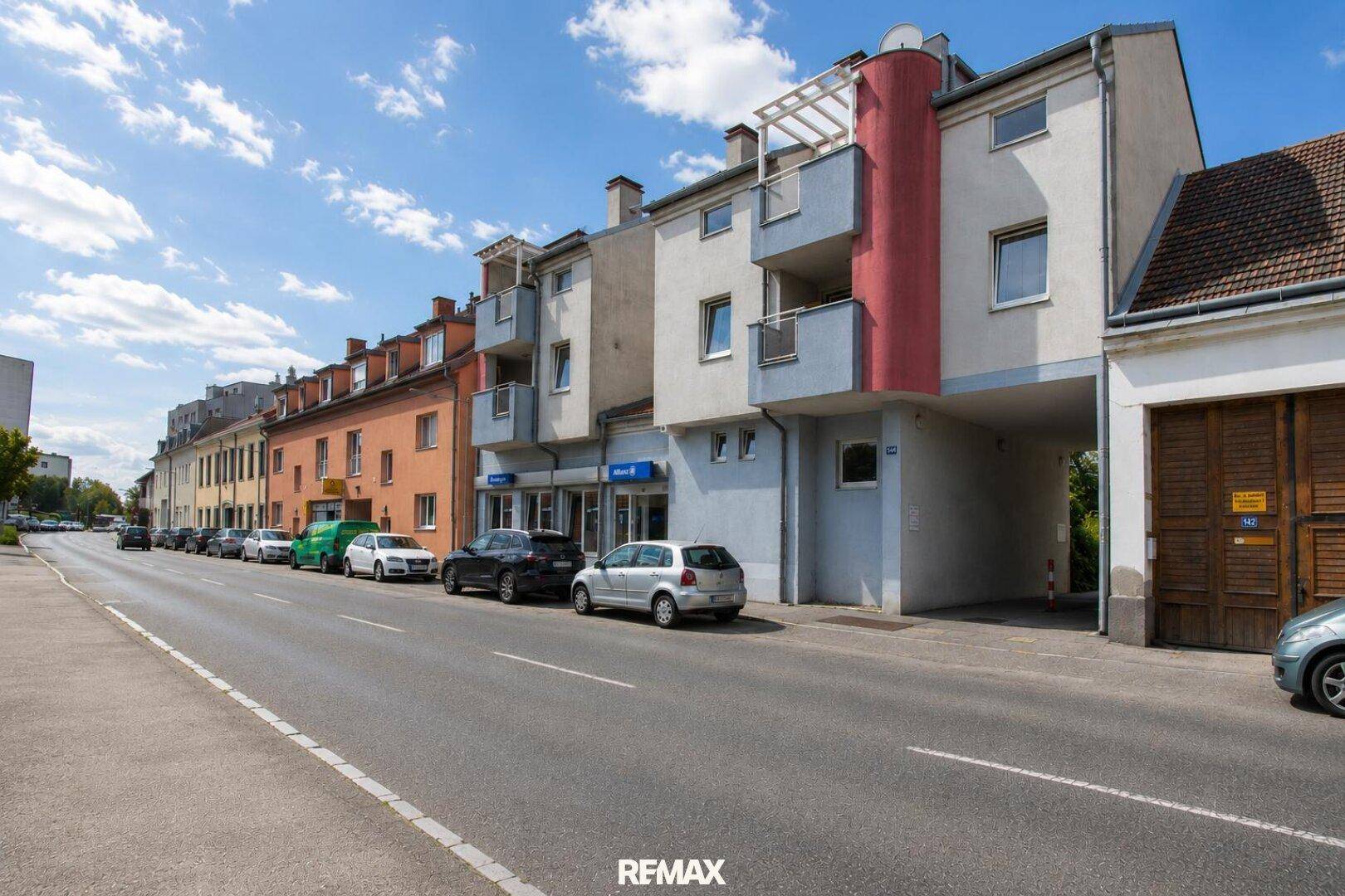 Wohngebäude an einer Straße mit parkenden Autos und Balkonen unter blauem Himmel.