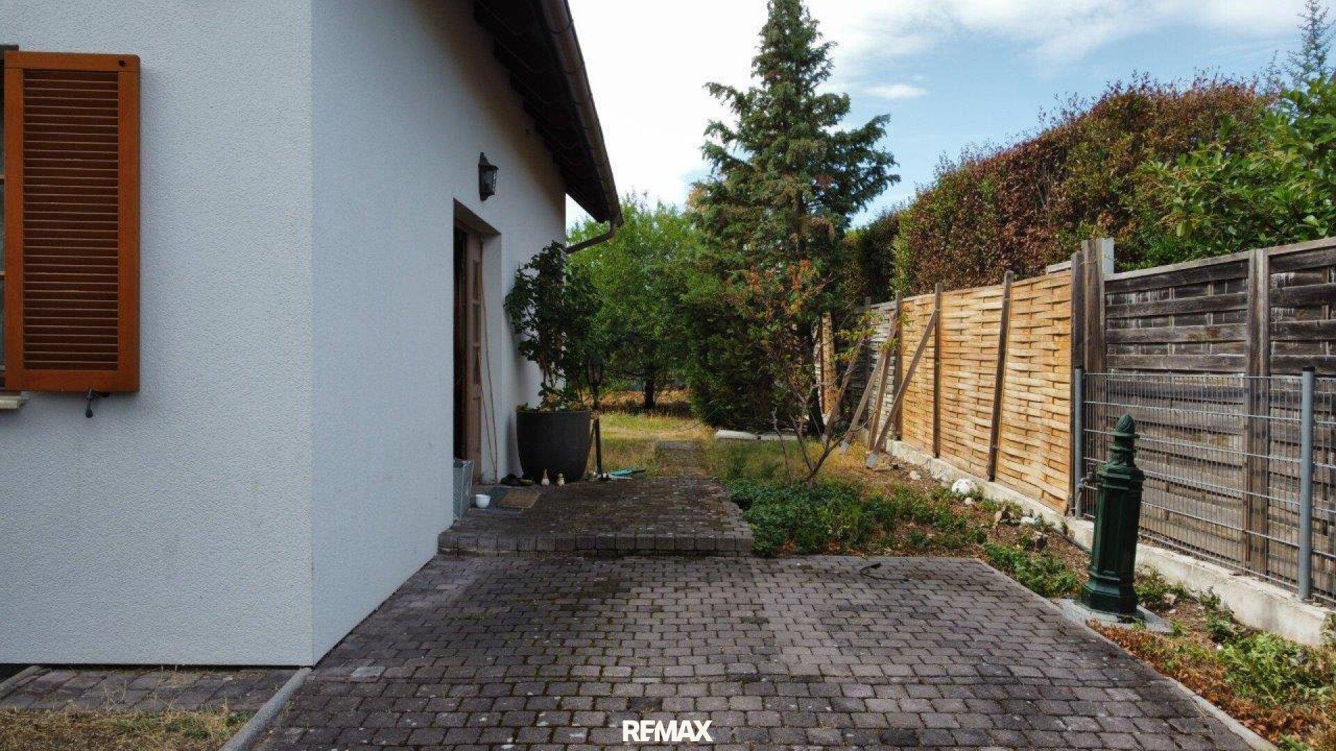 Gepflasterter Weg entlang der Hausseite mit Holzzaun und grüner Bepflanzung im Gartenbereich.