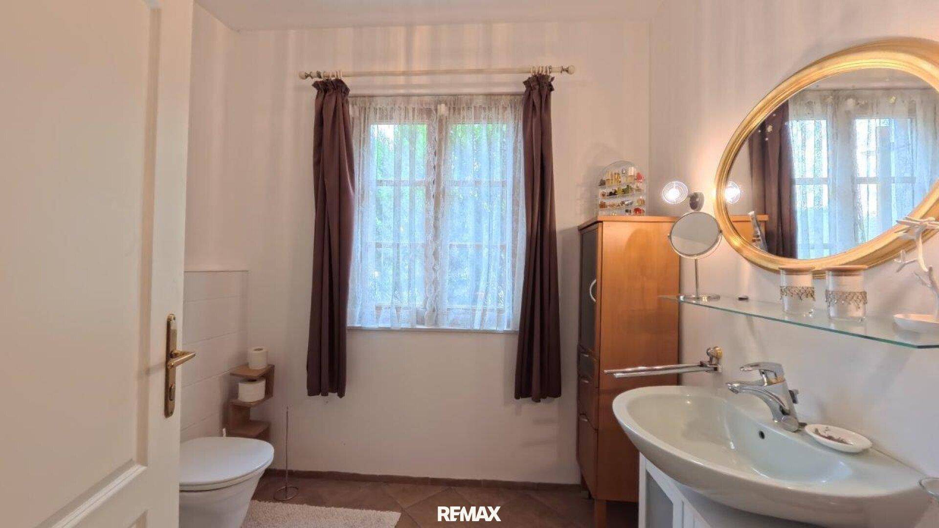 Badezimmer mit Fenster, Waschtisch und ovalem Spiegel in klassischem Stil.