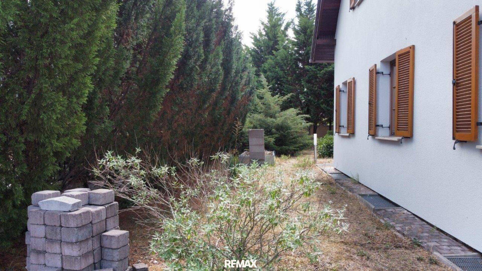 Seitenansicht des Hauses mit hohen Nadelbäumen und einem Bereich für Gartenarbeiten.