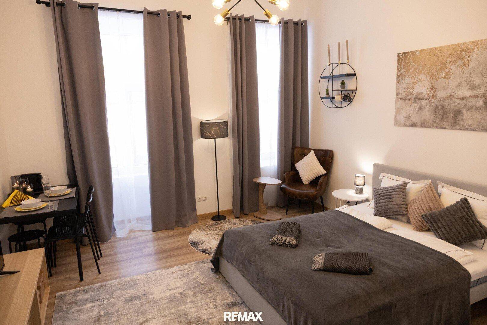 Helles Schlafzimmer mit Esstisch, Doppelbett und hohen Fenstern, ideal für ein Studio-Apartment.