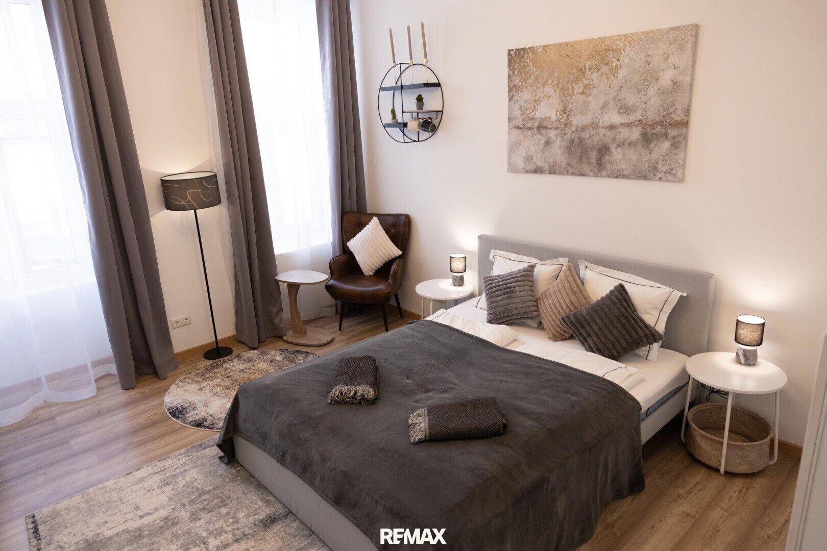 Schlafzimmer mit bequemen Bett, stilvollem Sessel und hohen Fenstern, die den Raum erhellen.