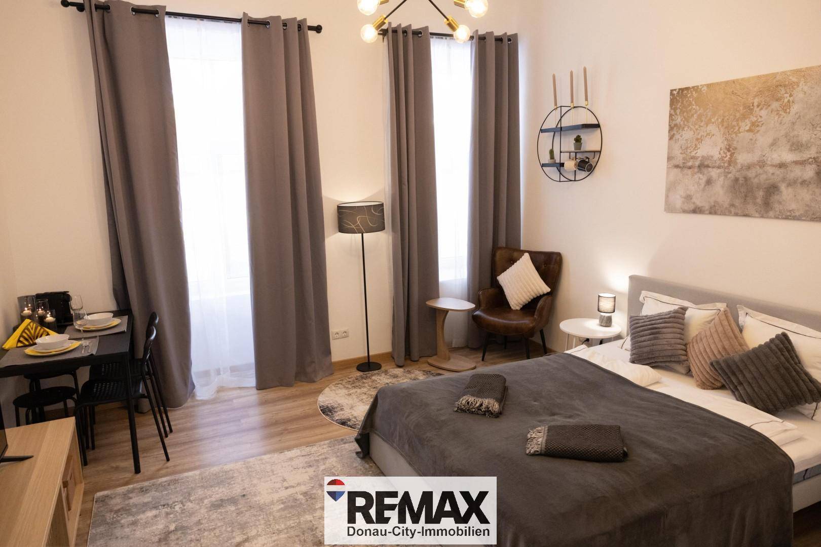 Funktionales Schlafzimmer mit Bett, kleinem Esstisch und viel Tageslicht durch große Fenster.