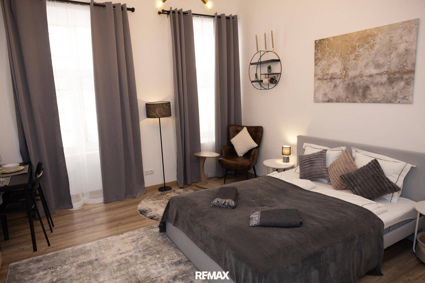 Gemütliches Schlafzimmer mit Doppelbett, Sessel und hohen Fenstern für viel Tageslicht.