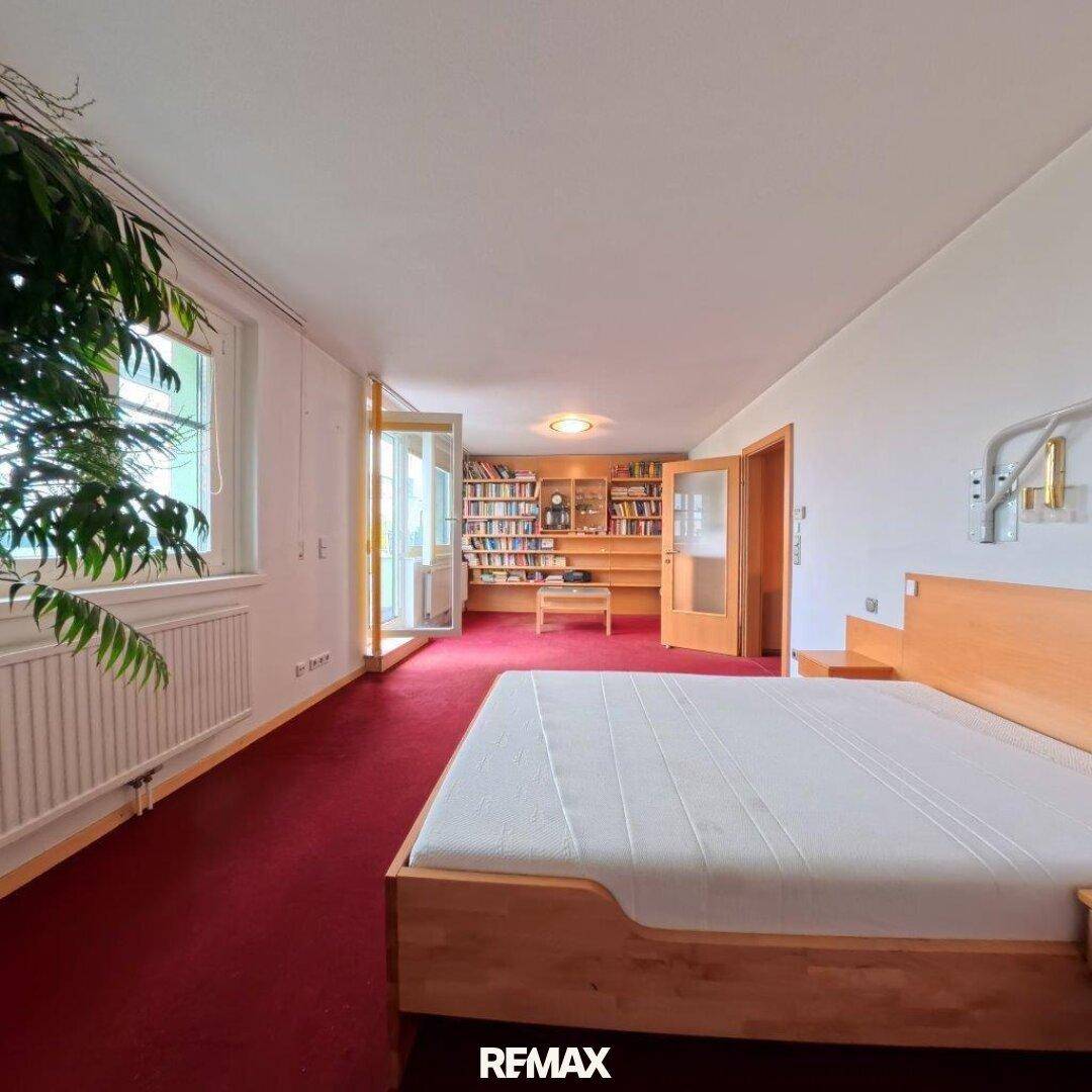 Helles Schlafzimmer mit rotem Teppichboden, Holzbett und Blick in ein angrenzendes Arbeitszimmer.