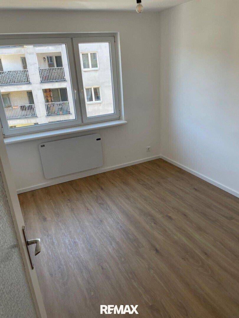 Leerer Raum mit Laminatboden, weißen Wänden und einem Fenster, das den Blick auf die umliegenden Gebäude freigibt.