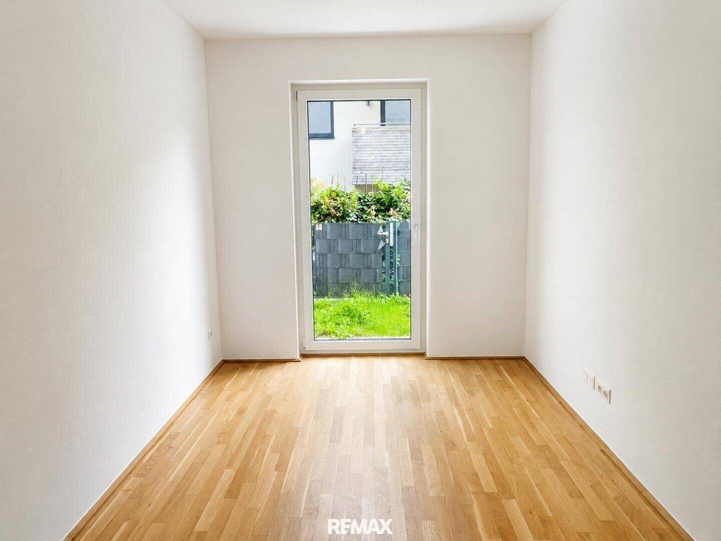 Helles Schlafzimmer mit Holzboden und Fenstertür, die den Blick in den kleinen Garten ermöglicht.