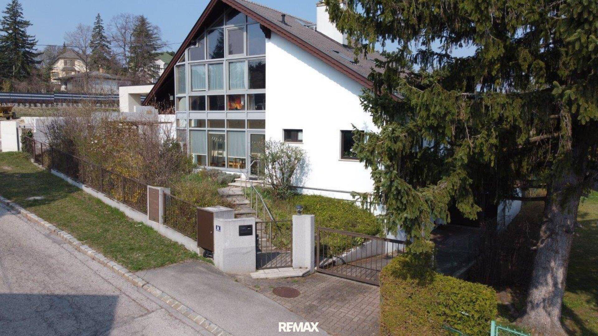 Moderne Hausfassade mit großer Glasfront und Satteldach, umgeben von einem gepflegten Vorgarten und Auffahrt.