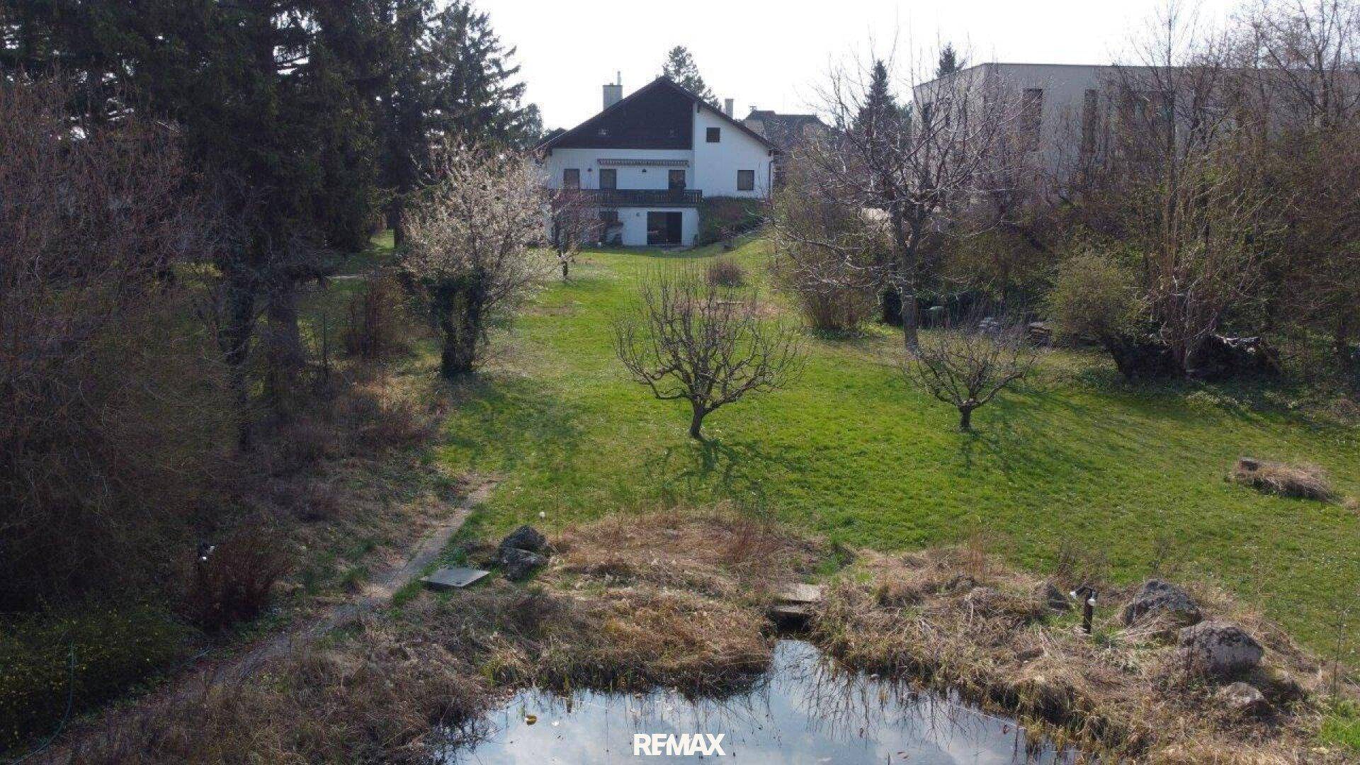 Blick vom Garten über den Teich auf das Wohnhaus, eingebettet in eine grüne Hügellandschaft.