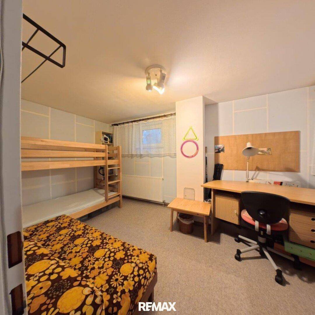 Kinderzimmer mit Etagenbett, Schreibtisch und gemustertem Teppichboden, gut beleuchtet.