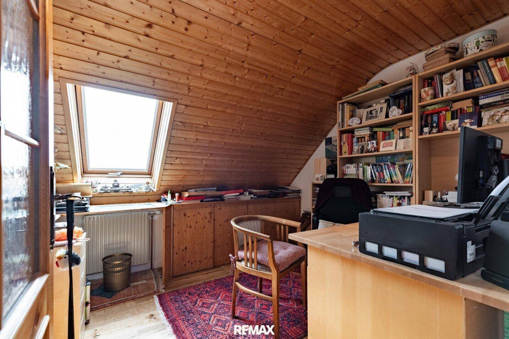 Helles Arbeitszimmer im Dachgeschoss mit Holzdecke, Schreibtisch, Bücherregalen und Dachfenster.