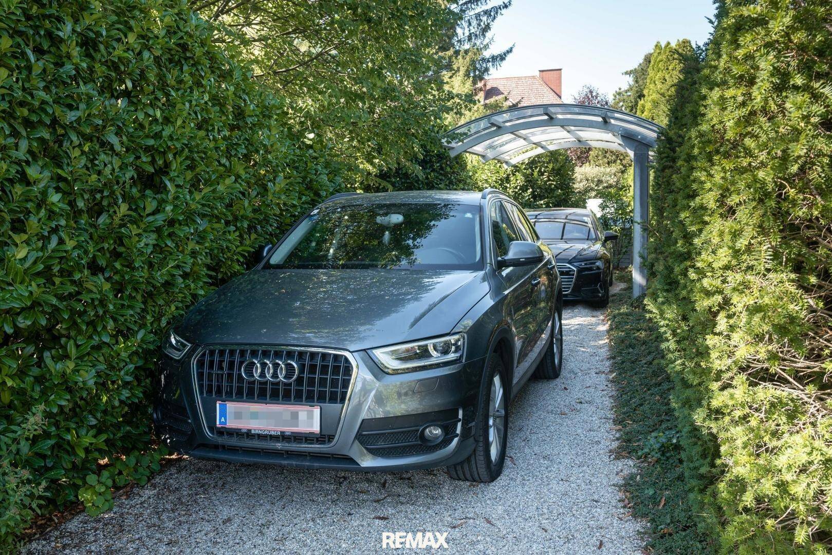 Einfahrt mit zwei Autos unter einem Carport, umgeben von hohen Hecken.