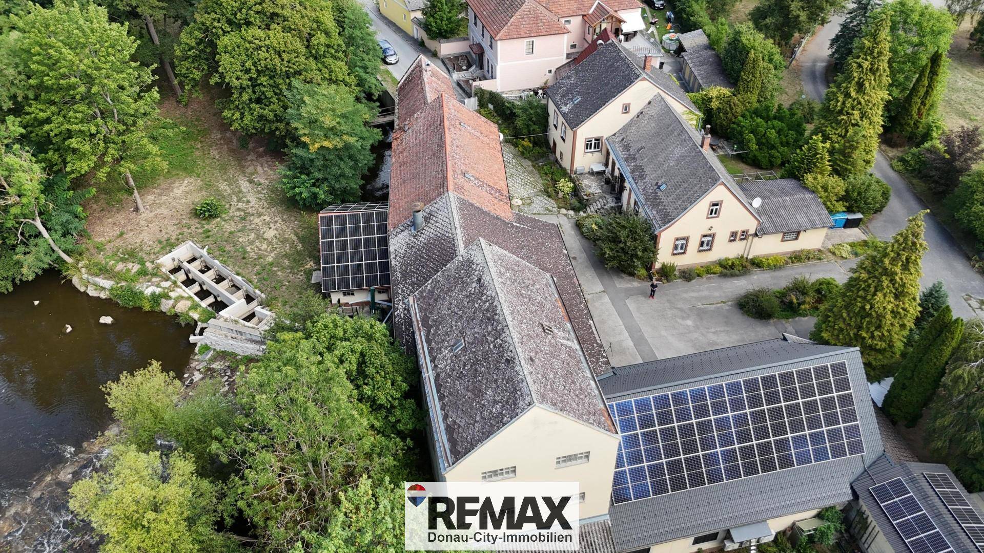 Luftaufnahme des Anwesens mit mehreren Gebäuden, Solaranlagen auf den Dächern und angrenzendem Fluss.