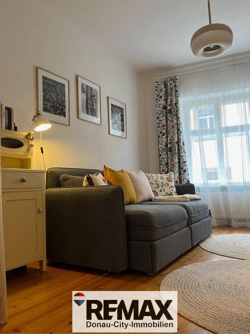Helles Schlafzimmer mit Schlafsofa, Holzboden und gemusterten Vorhängen am Fenster.