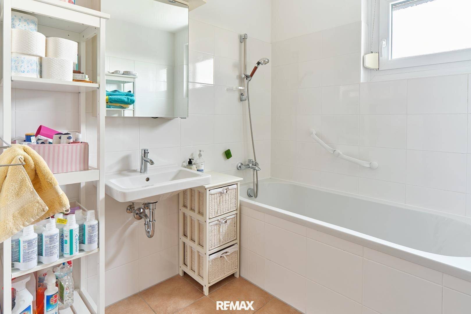 Helles Badezimmer mit Badewanne, Waschbecken und weißen Fliesen, ausgestattet mit einem Fenster.