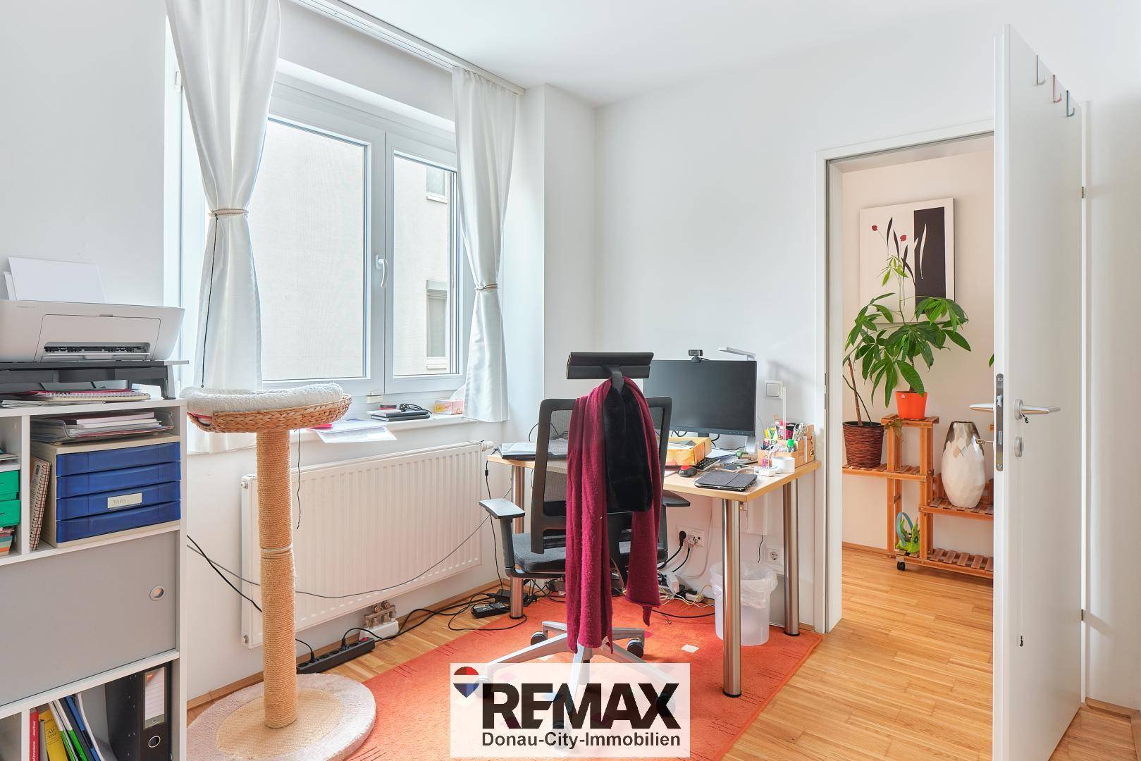 Helles Arbeitszimmer mit Schreibtisch, Fenster und Zugang zu einem weiteren Raum.