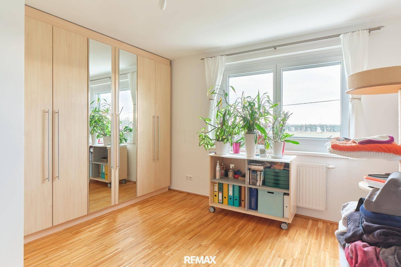 Schlafzimmer mit großen Spiegelschränken, Holzboden und Fenster mit Pflanzen.
