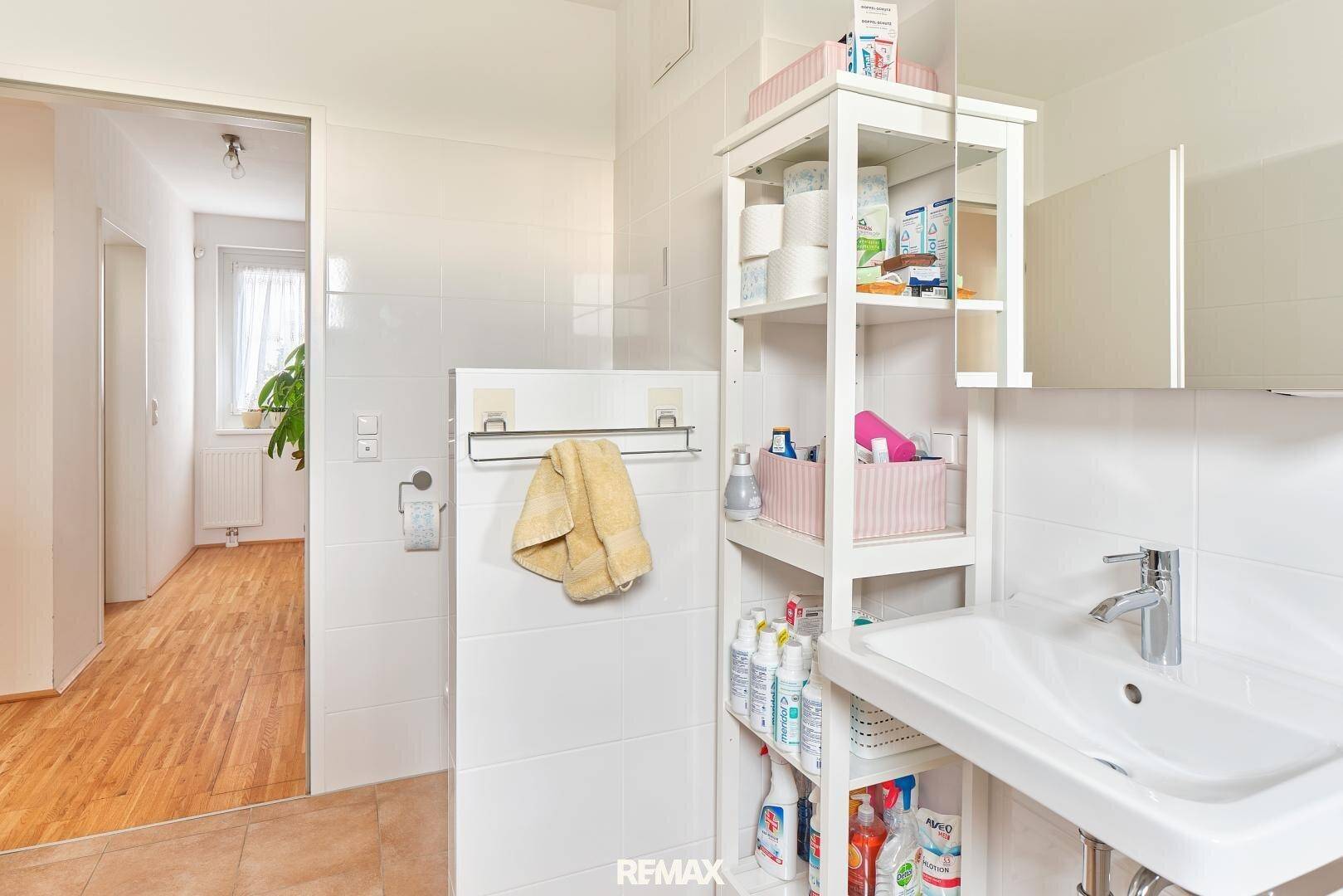Badezimmer mit Waschbecken, Regal und weißen Fliesen, mit Blick in den Flur.
