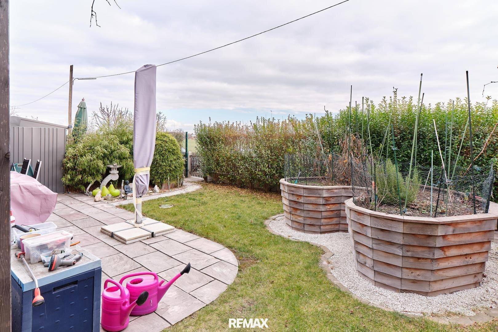 Garten mit gepflasterter Terrasse, Hochbeeten und einem Sonnenschirm.