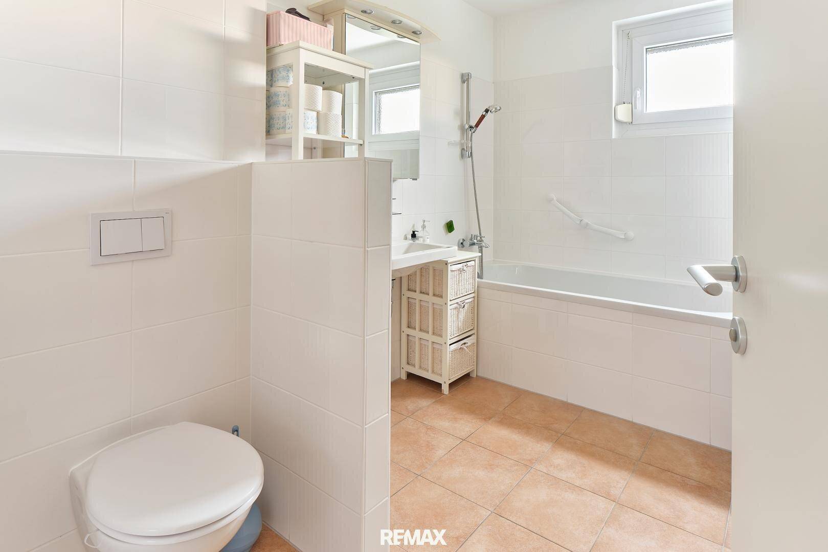 Modernes Badezimmer mit Badewanne, WC und weißen Fliesen, gut beleuchtet durch ein Fenster.