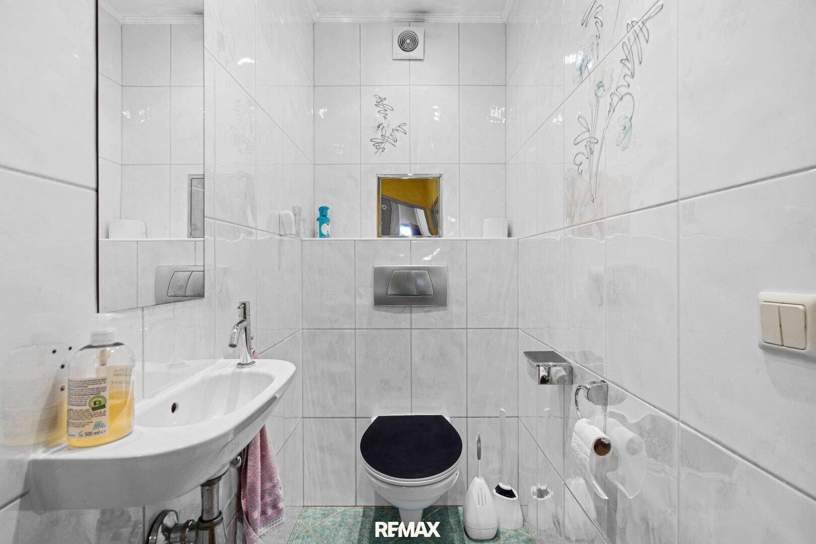 Separates WC mit wandhängender Toilette, kleinem Waschbecken und weißen Fliesen.