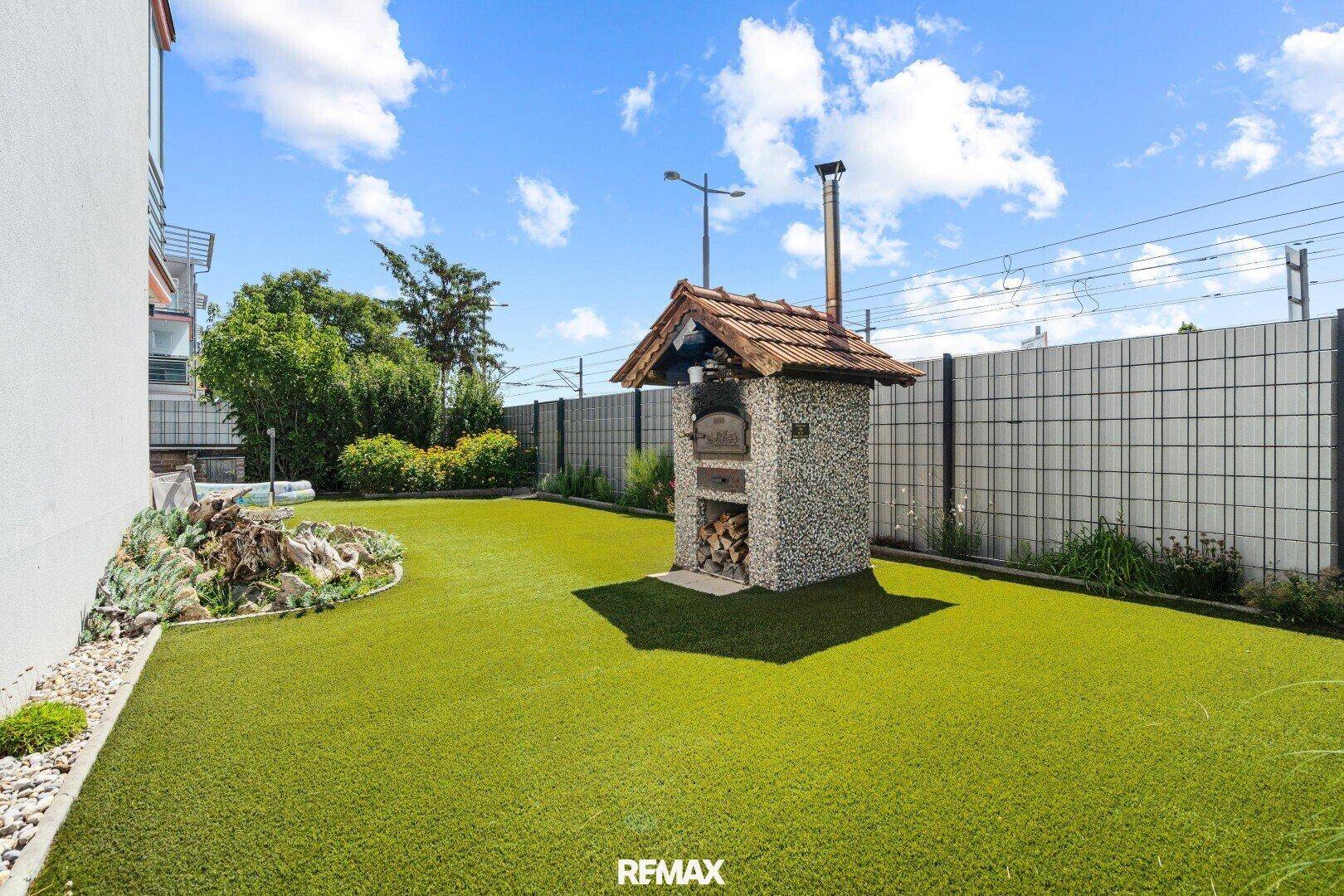 Gepflegter Gartenbereich mit Kunstrasen, einem traditionellen Pizzaofen und Blick auf die Umgebung.